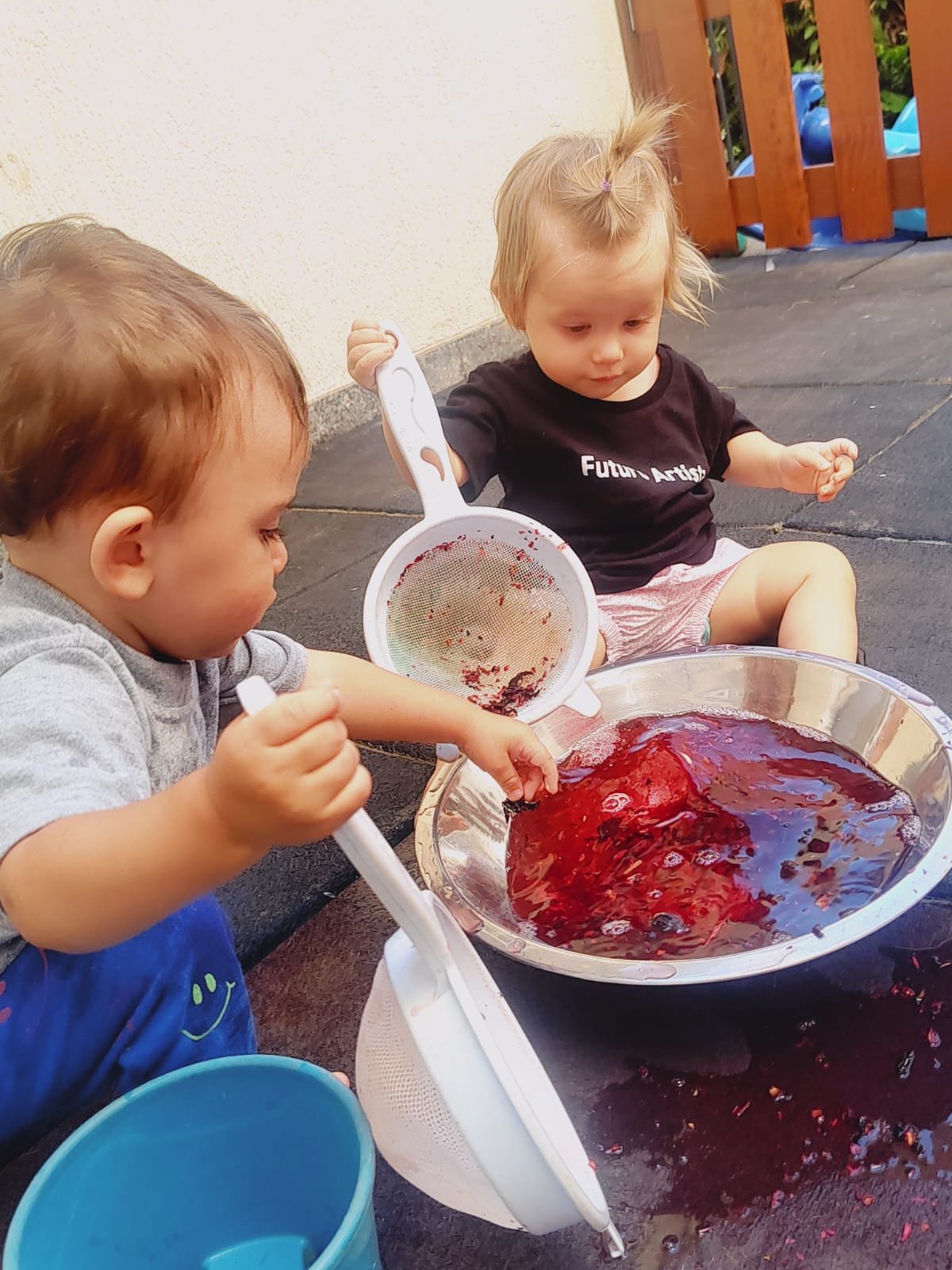 Dois bebês brincando com peneiras e uma bacia de água colorida
