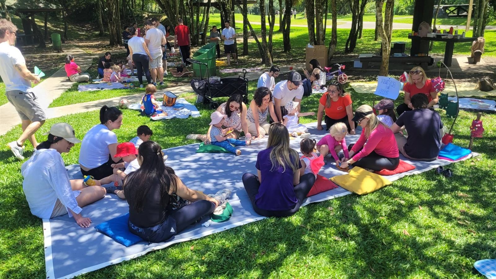 Famílias, bebês e educadoras brincando juntos sobre uma lona no parque